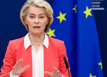 Ursula von der Leyen, pink suit, white collar shirt, blonde hair, black microphone, EU flag, stars