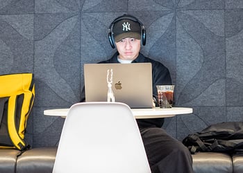 Man wearing headphones and baseball cap working on laptop at cafe table with backpack and iced coffee