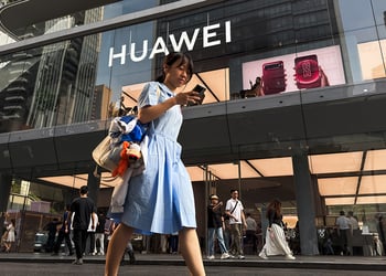 A pedestrian looks at a smartphone while walking past a Huawei retail store displaying an illuminated Huawei sign and smartphone advertisements 