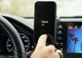 uber app open on black smartphone screen, car screen, blue sky and palm tree, driving wheel
