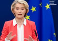 Ursula von der Leyen, pink suit, white collar shirt, blonde hair, black microphone, EU flag, stars