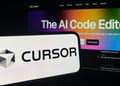 Cursor logo on phone, background AI coding agent written on computer screen.