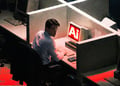 Man sitting in front of PC saying "AI", and looking back, mischief in his face, in dark office